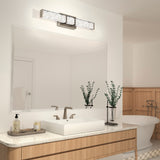 Modern bathroom with a large mirror above a light wood vanity, a rectangular sink, and elegant faucet. Two glass vases with leafy green stems and a soap dispenser are on the countertop. A bathtub is visible in the background.
