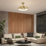 Modern living room with a light beige sectional sofa, green and black pillows, a round black marble coffee table, wood-paneled wall, large plant, and a gold ceiling light fixture. Black blinds cover a window on the right.