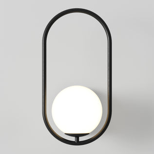 The Artika Bloom LED Sconce 3 CCT features a sleek black oval frame with a glowing white spherical bulb elegantly suspended at the center, creating a modern look against any light gray wall.
