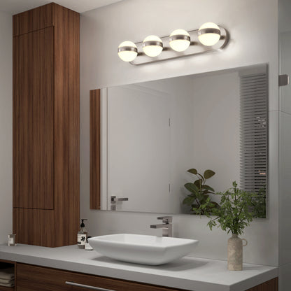 A modern bathroom features a large mirror, wooden cabinets, a vessel sink, and the Artika Baldric LED modern bathroom vanity light above the mirror. A potted plant and soap dispenser decorate the countertop; blinds cover the window on the right.