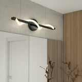 A modern black wall sconce with a twisted LED light mounted above a large mirror in a minimal bathroom with light wood paneling and a white door. Brown decorative branches are visible in the foreground.