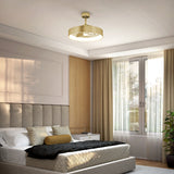 A modern bedroom with a beige upholstered bed, pillows, a black throw, a nightstand, large window with sheer curtains, and a gold ceiling light fixture. Natural light fills the room, and a plant is in the corner.