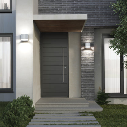 Modern home entryway with a dark gray door, brick and concrete walls, small porch roof, window with white curtains, landscaped greenery, and two Artika Valor Square LED indoor-outdoor wall lights.