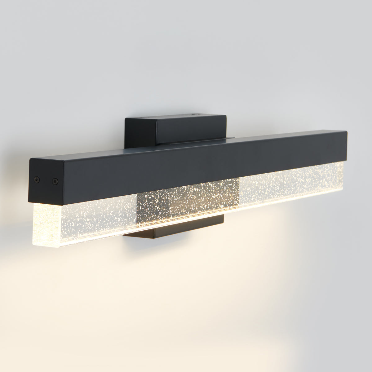 A modern wall-mounted light fixture with a rectangular black frame and a clear section featuring small bubble-like patterns, emitting a soft, warm glow against a light-colored wall.