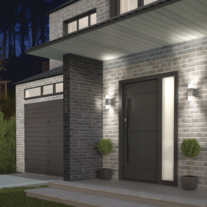 Modern house exterior at dusk with gray brick walls, dark garage door, large black front door, two Artika Lennox indoor-outdoor LED wall lights, two potted topiary plants, and house number 635 visible on the brick column.