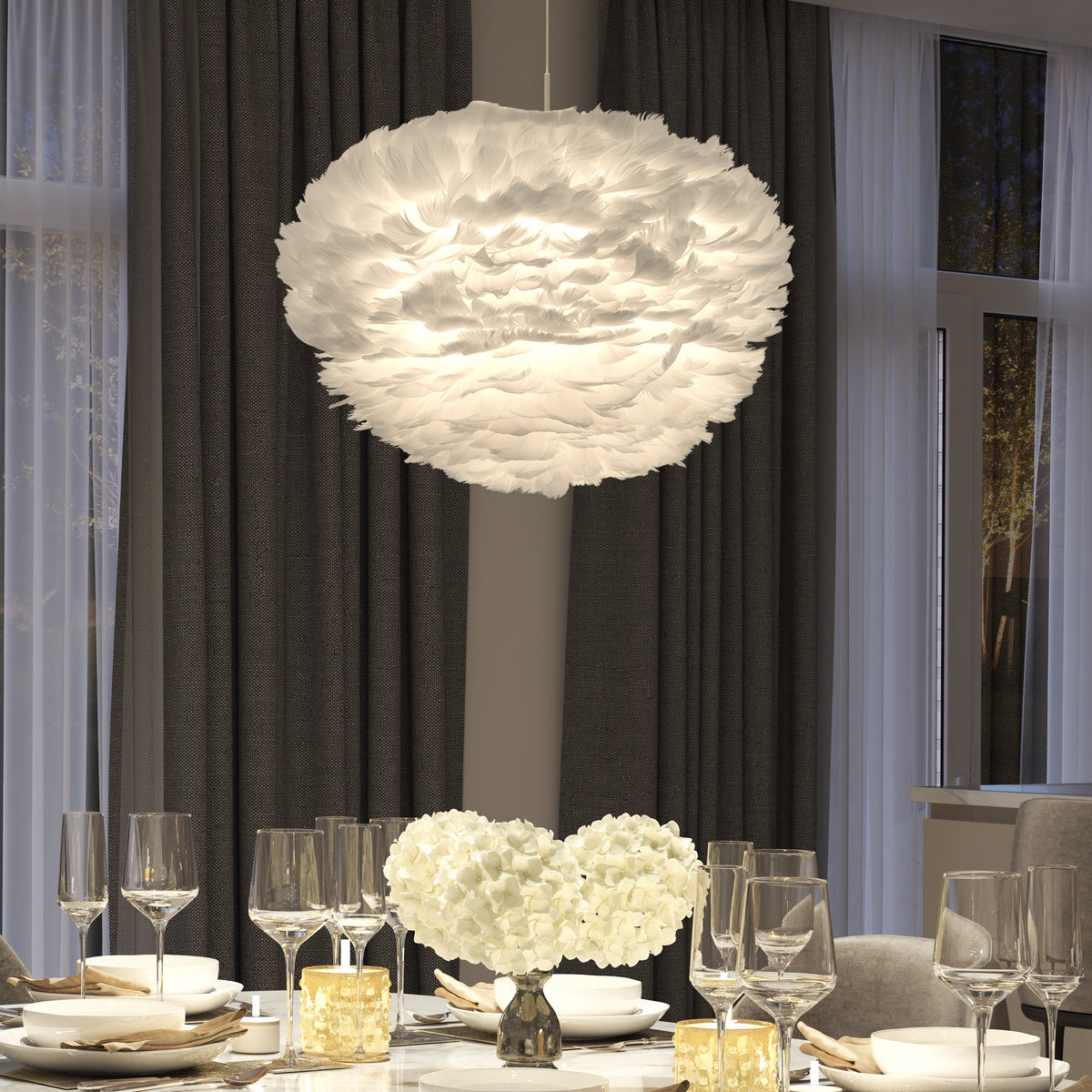 A modern dining room with a fluffy, white feather pendant light hanging above a table set with wine glasses, white plates, and white flower centerpieces, against dark curtains and large windows.