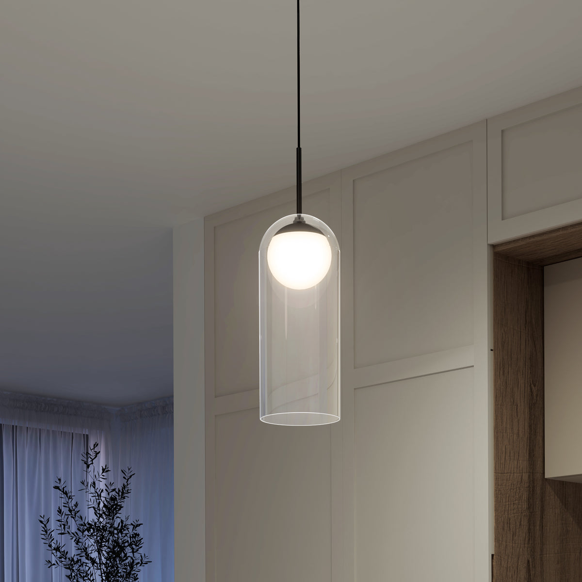A modern pendant light with a clear glass cylindrical shade and a glowing white bulb hangs from the ceiling in a minimalist room with neutral cabinets and a plant in the corner.