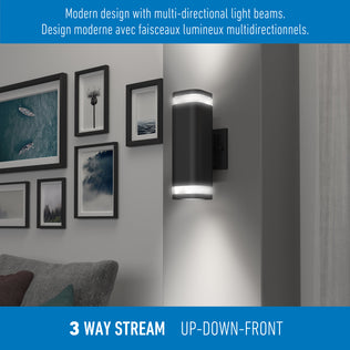 The Artika Stark modern LED indoor-outdoor light with wi-fi features a sleek black design, casting light up, down, and forward to highlight artwork above a gray sofa. Bilingual English and French text describes its multi-directional style.