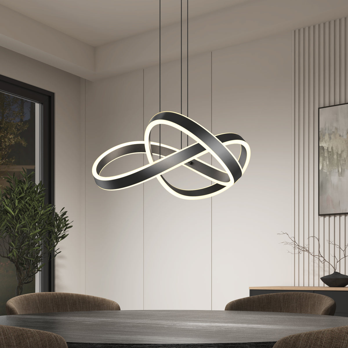 A modern dining room with a round table and dark chairs features an abstract, intertwined LED chandelier hanging from the ceiling. The room has minimal decor, with a potted plant and a framed artwork on the wall.