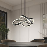 A modern dining room with a round table and dark chairs features an abstract, intertwined LED chandelier hanging from the ceiling. The room has minimal decor, with a potted plant and a framed artwork on the wall.