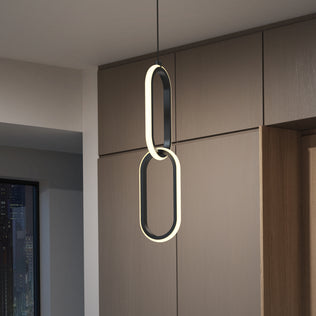 The Catena LED pendant light 5 CCT by Artika, featuring two vertically connected oval loops, hangs from the ceiling in a contemporary room with wood panel walls and a city view through the window.