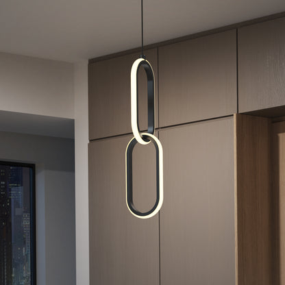 The Catena LED pendant light 5 CCT by Artika, featuring two vertically connected oval loops, hangs from the ceiling in a contemporary room with wood panel walls and a city view through the window.