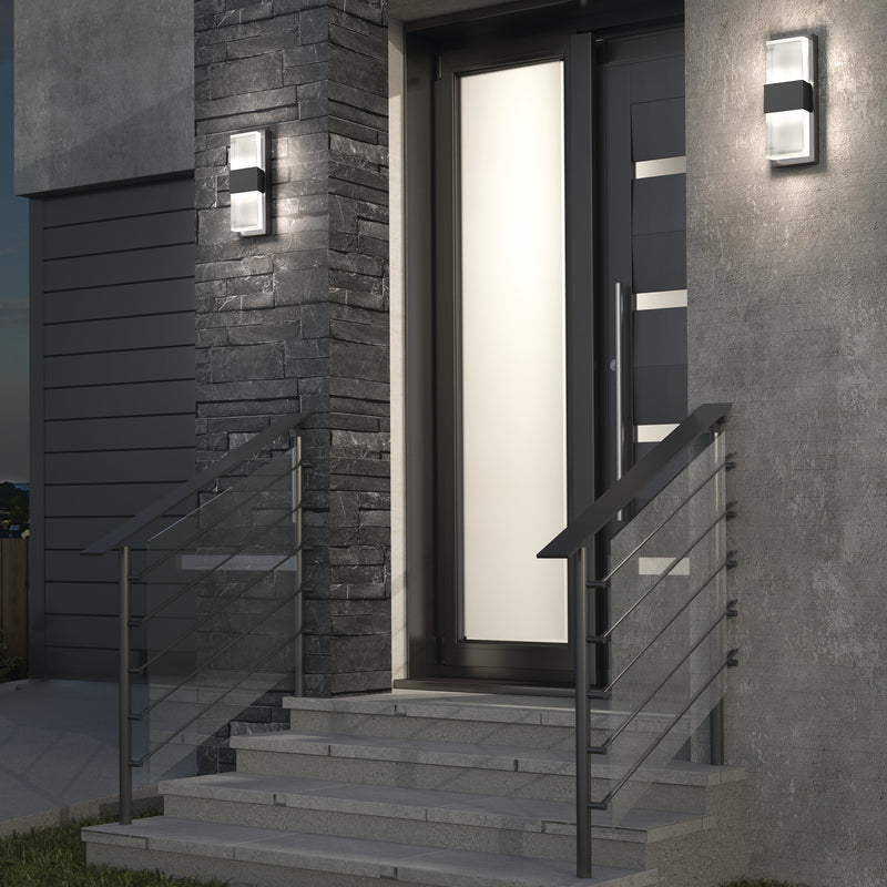 Modern house entrance with gray stone and concrete walls, black metal railings, and Artika Mazza Frosted indoor-outdoor LED wall lights flanking a frosted glass front door. Steps lead up to the entrance.