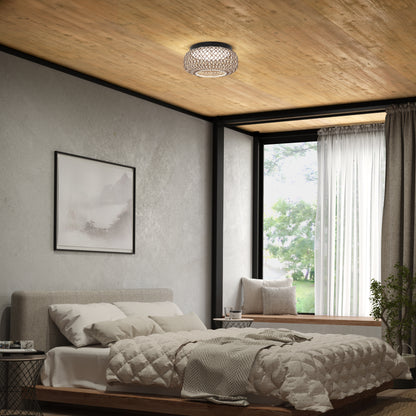 A cozy bedroom with a beige bed, quilted bedding, a large window with curtains, a potted plant, framed landscape art on the wall, and an Artika Benson 15 in. LED flush mount light 5 CCT on the wooden ceiling.