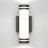 A modern wall-mounted cylindrical light fixture with black accents at the top and bottom, emitting a soft white glow against a plain light gray wall.