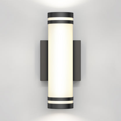 A modern wall-mounted cylindrical light fixture with black accents at the top and bottom, emitting a soft white glow against a plain light gray wall.