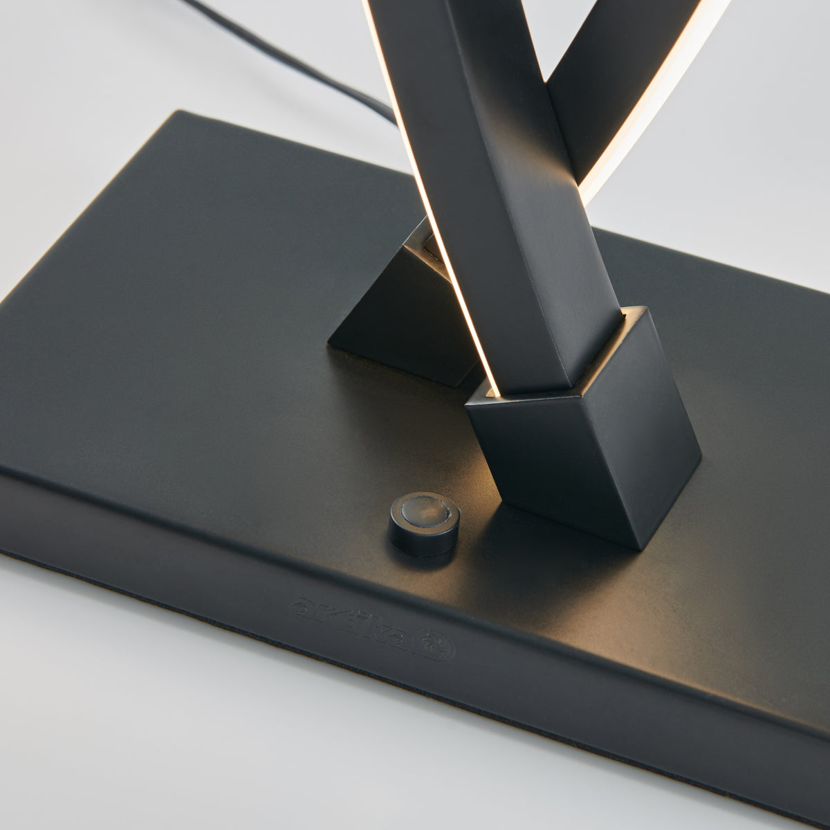 Close-up of a modern lamp base with intersecting black metal bars and an integrated LED light strip, featuring a small round button and subtle brand text on the rectangular base.