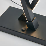 Close-up of a modern lamp base with intersecting black metal bars and an integrated LED light strip, featuring a small round button and subtle brand text on the rectangular base.