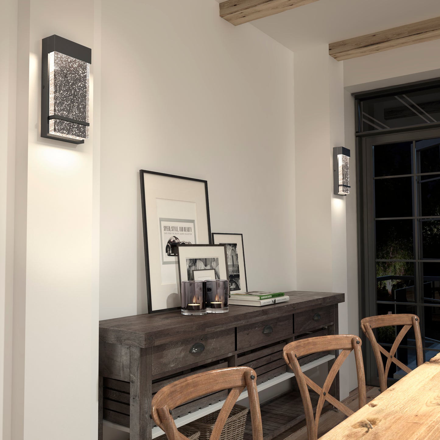 A rustic dining room features a wooden table, chairs, and a sideboard with framed art and candles. Two Artika Skyler indoor-outdoor LED wall lights softly illuminate the white walls above the decor.