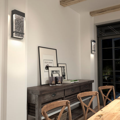 A rustic dining room features a wooden table, chairs, and a sideboard with framed art and candles. Two Artika Skyler indoor-outdoor LED wall lights softly illuminate the white walls above the decor.