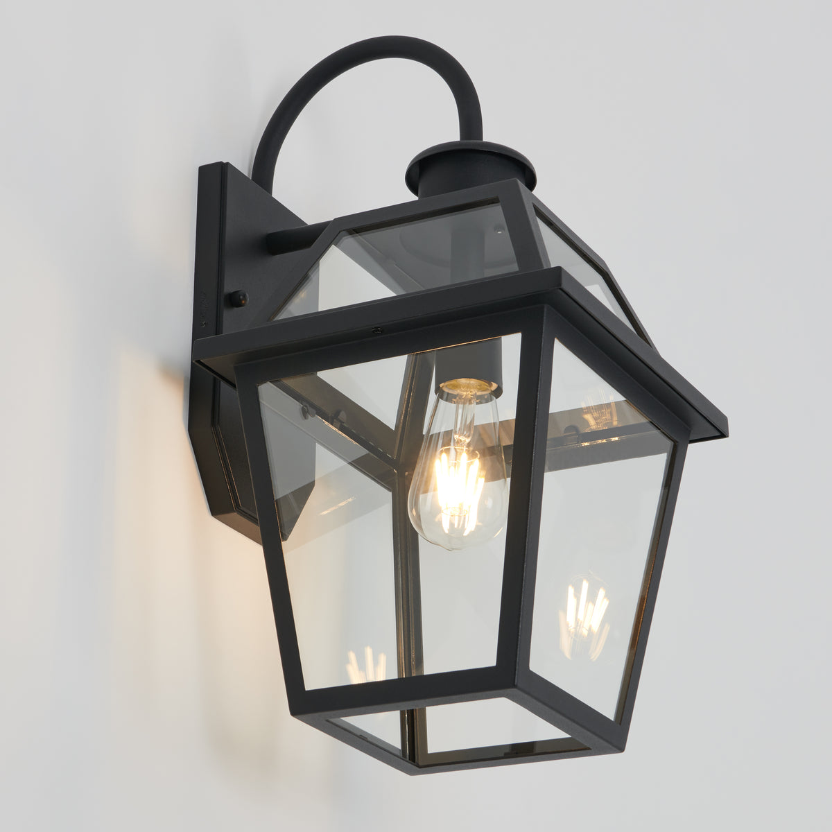 The Artika Colonial outdoor lantern wall light features a black metal and glass design with a visible LED bulb, classic geometric lines, and a curved top arm, shown mounted on a white wall.