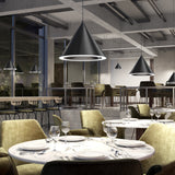 A modern restaurant interior with round marble tables, green velvet chairs, wine glasses, and large black pendant lights. The space has floor-to-ceiling windows and an elevated seating area in the background.