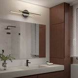 Modern bathroom with a large mirror, double sink, and black fixtures. A horizontal light fixture is above the mirror. There are wooden cabinets, a small plant, a soap dish, and a window with white blinds.