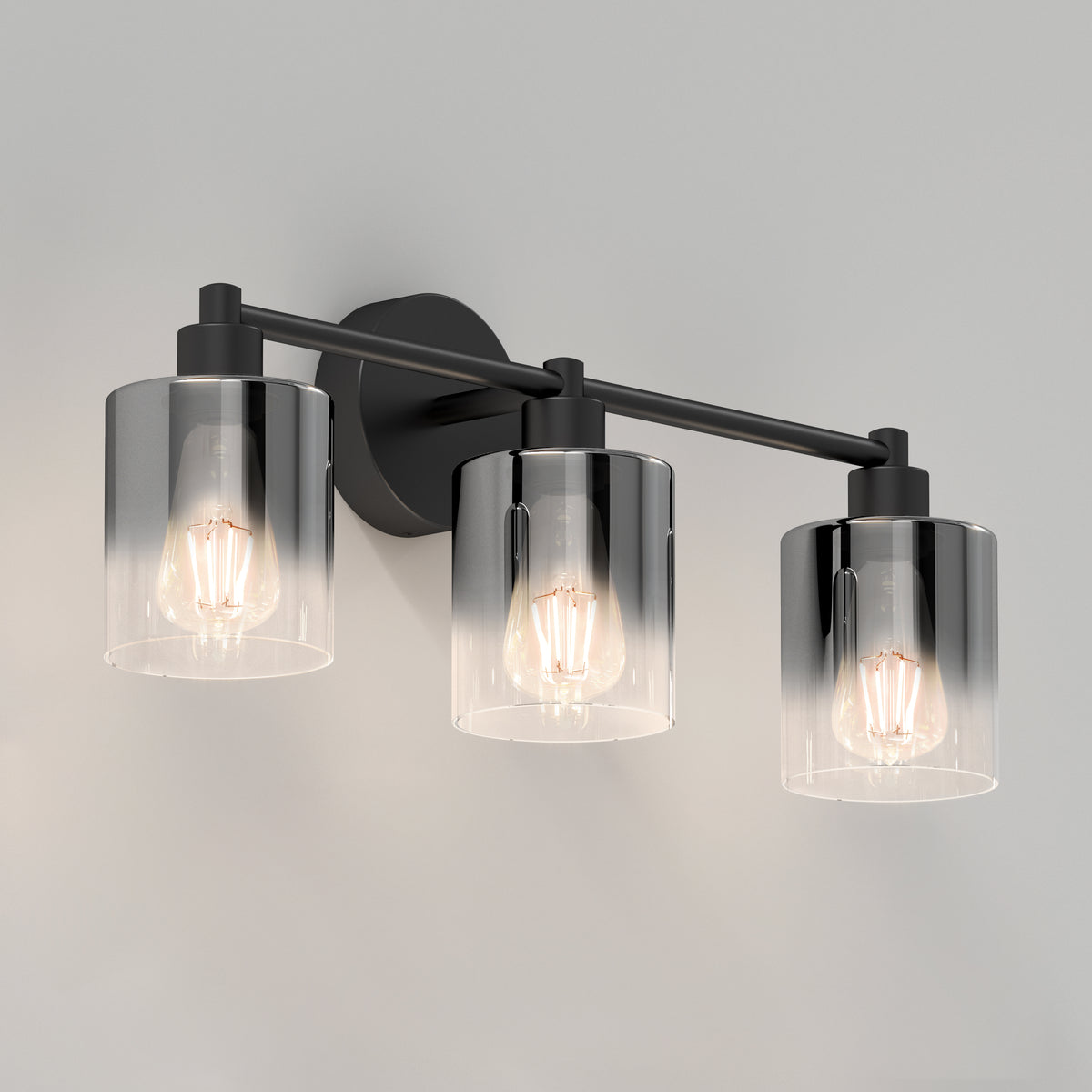 A modern wall-mounted light fixture with three exposed bulbs, each covered by a cylindrical clear glass shade, set on a horizontal black metal bar against a white background.