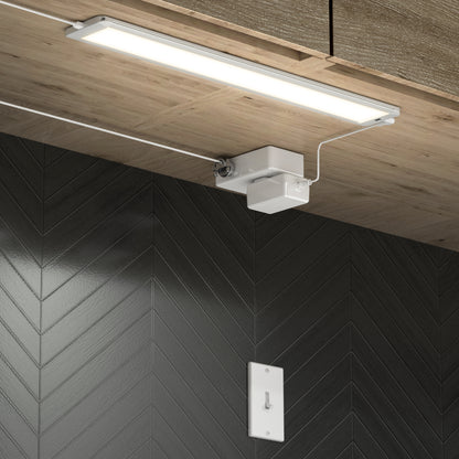 The Artika Maestro under cabinet LED light 3CCT, a slim rectangular fixture, is mounted beneath a wooden cabinet to brighten a dark chevron-patterned tile backsplash. A switch plate appears on the wall below the light.