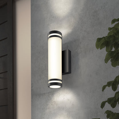 A modern cylindrical wall sconce with a black frame and frosted glass emits light both upward and downward against a gray textured wall, with green leafy plants partially visible on the right side.