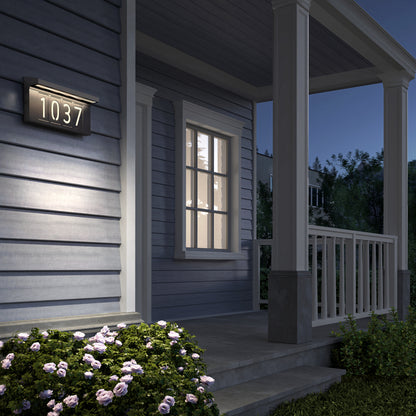 A gray-sided house with a front porch is lit by the Artika Bowen solar LED outdoor address wall light 5 CCT, displaying 1037. Pink flowers and green shrubs line the steps in the garden at twilight.