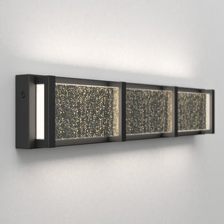 The Artika Subway Essence 27-in Integrated LED Vanity Light Bar features a sleek black frame and three textured glass panels, casting a warm glow—ideal for bathroom lighting against a white wall.
