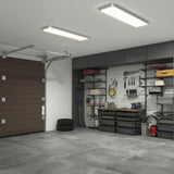 A clean, organized garage with gray shelves holding tools, storage bins, and equipment along one wall. A brown garage door is partially visible on the left, with stacked tires and gray cabinets overhead.