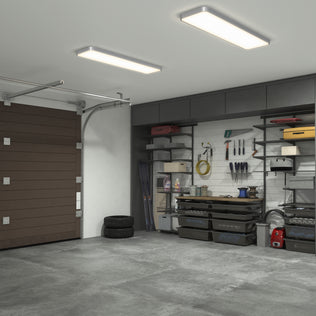 A clean, organized garage lit by the Artika Europa 48 LED flush mount light with remote. Wall-mounted shelves hold tools and supplies. Stacked tires sit on the left by a closed brown garage door, and the concrete floor is uncluttered.