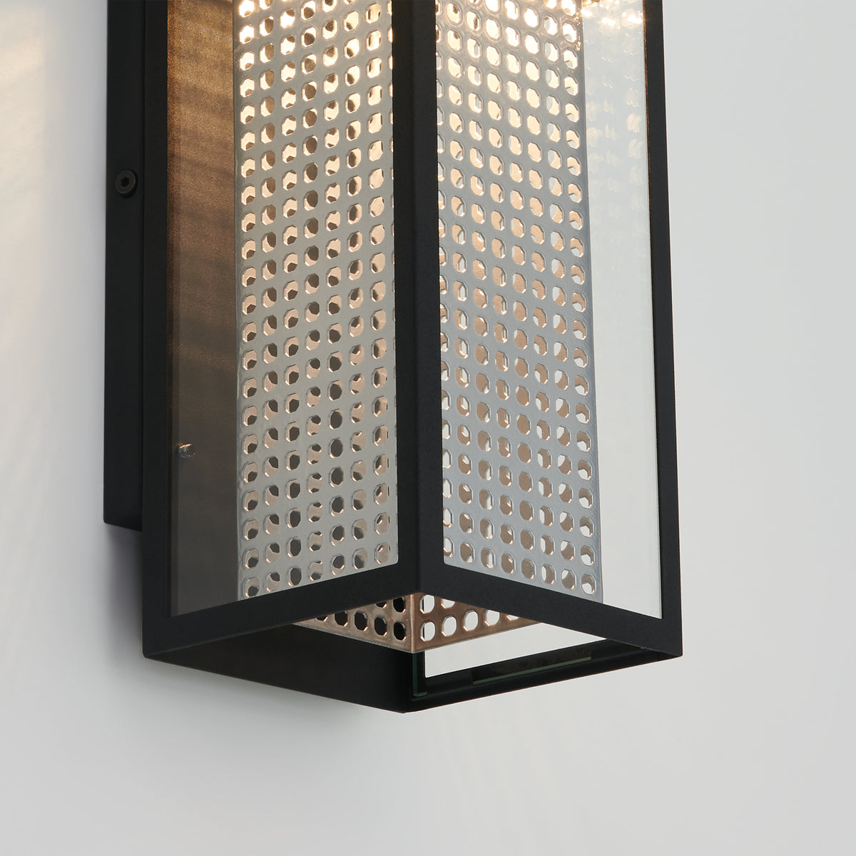 A modern wall sconce with a black metal frame and perforated metal panels, allowing light to shine through the small circular holes, mounted on a white wall.
