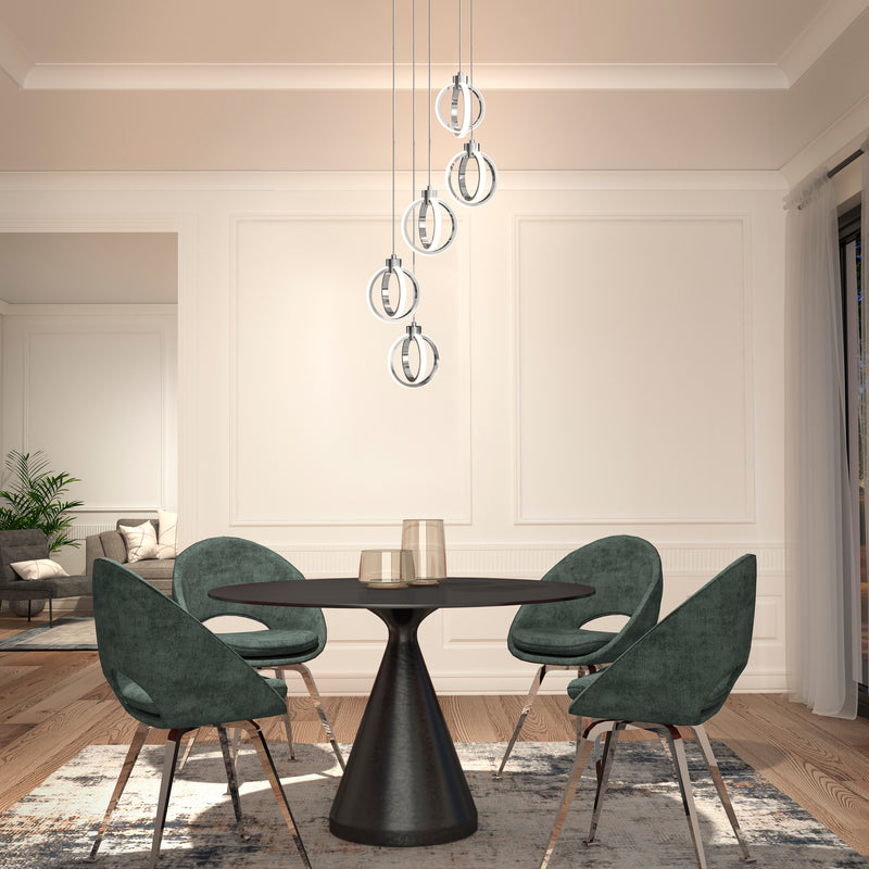 A modern dining room featuring a round black table, four green velvet chairs, the Artika Soho modern 5-light LED pendant, white paneled walls, wood flooring, a large window with sheer curtains, and a corner plant.