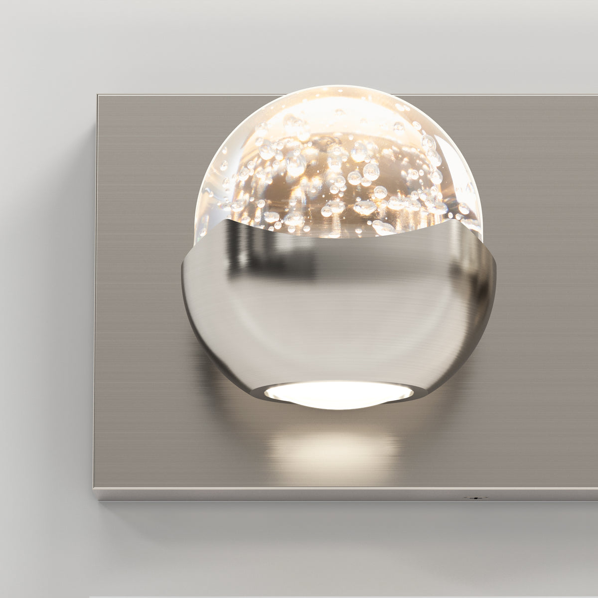 A modern wall sconce with a metallic base and a clear glass globe partially encased, featuring small bubbles inside the glass, emits a soft light against a light gray wall.