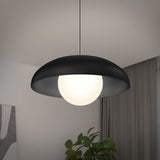 A modern pendant light with a black dome-shaped shade and a round white bulb hangs from the ceiling in a minimalist room with neutral walls, curtains, a plant, and a partially visible abstract wall art.