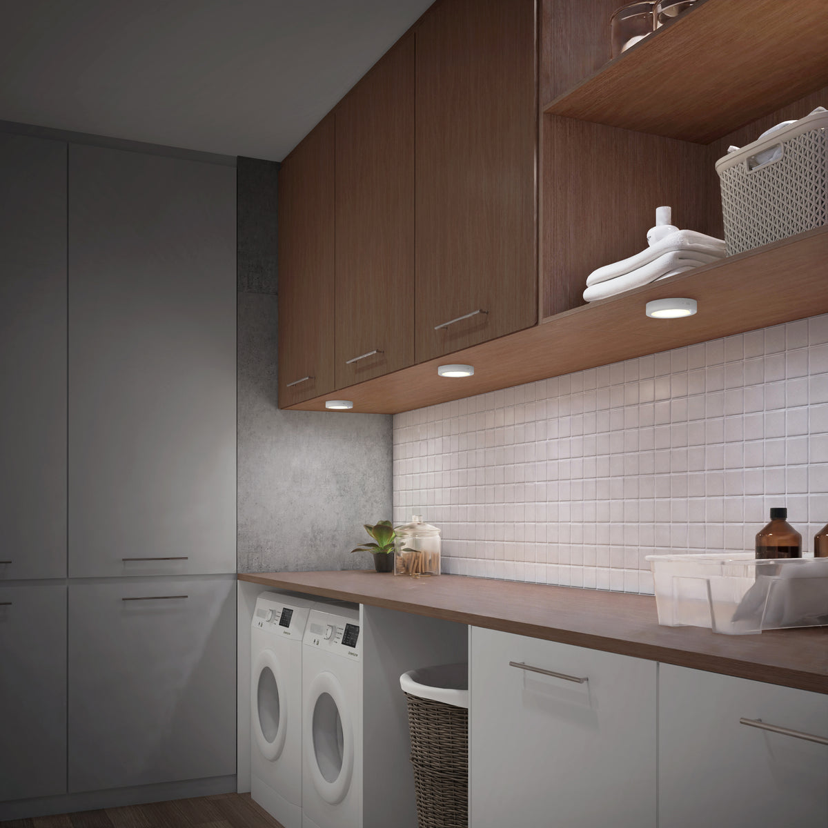 A modern laundry room with white cabinets, wood upper cabinets, a countertop, two front-loading washing machines, white tile backsplash, and baskets for storage. Under-cabinet lights illuminate the workspace.