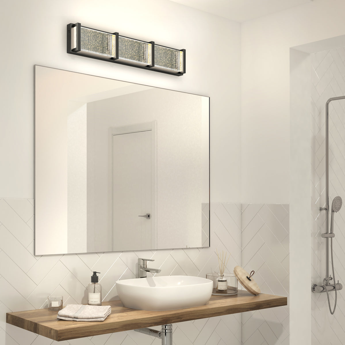 Modern bathroom with a large mirror, rectangular vessel sink on a wooden countertop, soap dispenser, rolled towel, reed diffuser, and a three-light wall fixture above the mirror; partial view of a shower on the right.