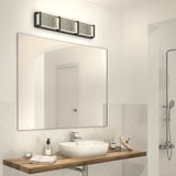 Modern bathroom with a large mirror, rectangular vessel sink on a wooden countertop, soap dispenser, rolled towel, reed diffuser, and a three-light wall fixture above the mirror; partial view of a shower on the right.