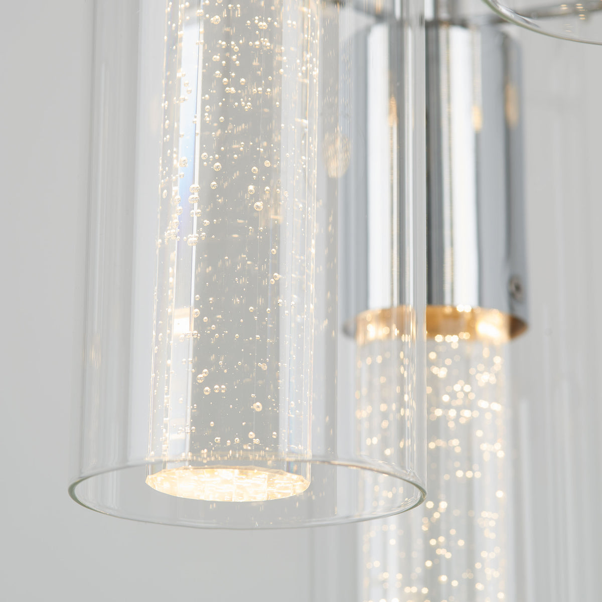 Close-up of modern cylindrical glass light fixtures with tiny bubbles inside, emitting a soft, warm glow. The fixtures have metallic accents and are suspended against a light, neutral background.