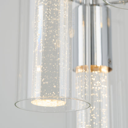 Close-up of the Artika Bubble Swirl 5-light pendant, featuring modern cylindrical glass shades with tiny bubbles and a sleek metallic finish, emitting a soft, warm glow in a contemporary design.