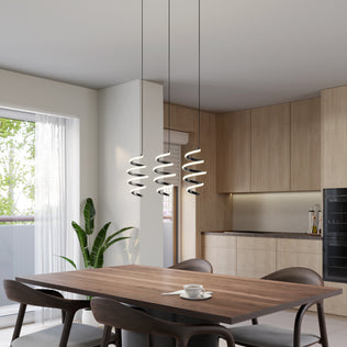 A modern kitchen and dining area features light wood cabinets, a dark wood dining table with four chairs, a potted plant, sheer-curtained window with greenery view, and Artika’s Chorus 3-light LED pendant light (5 CCT).