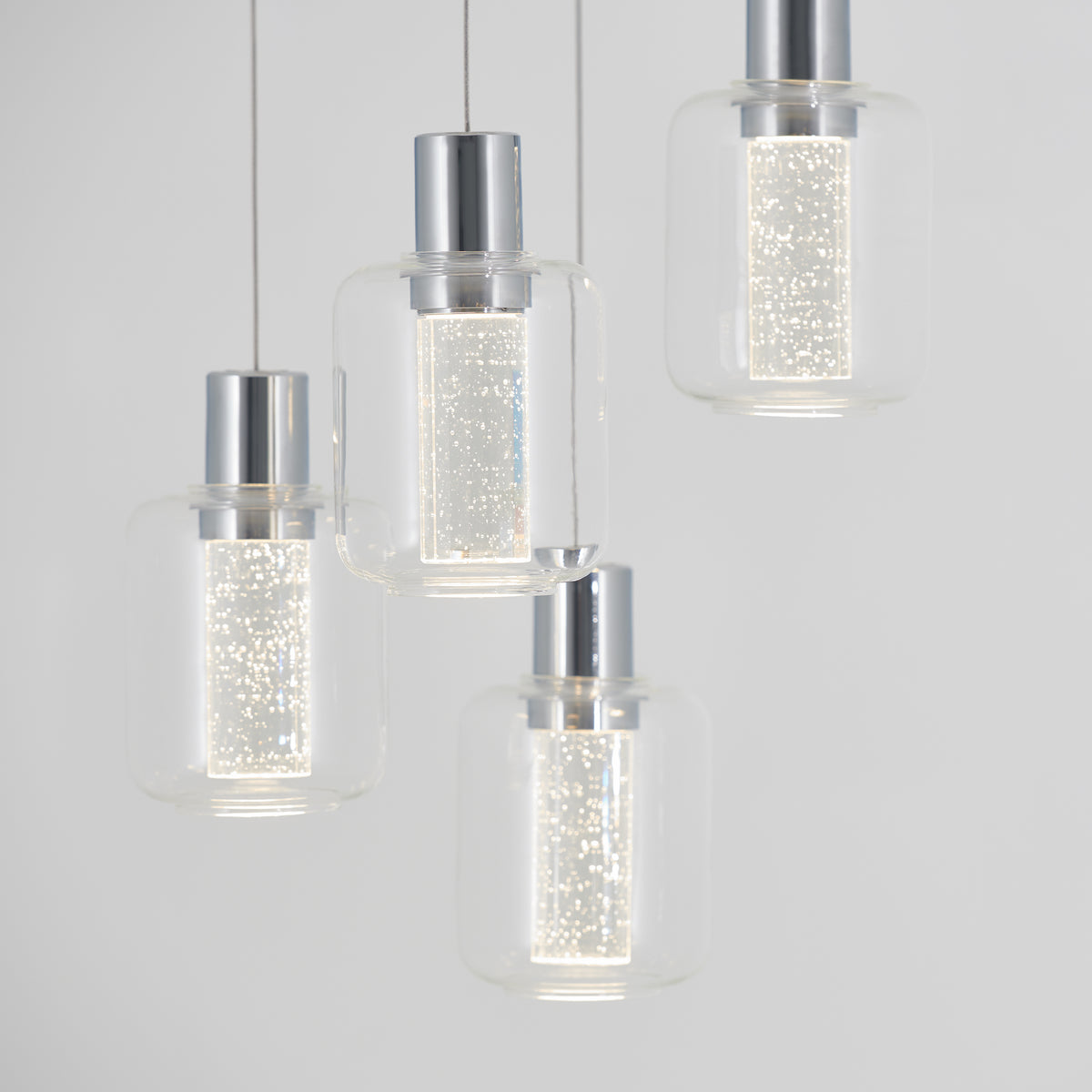 Four modern pendant lights with clear glass shades and metallic tops hang at different heights. The lights inside each shade emit a bright, sparkling glow, creating a contemporary and elegant look against a light background.