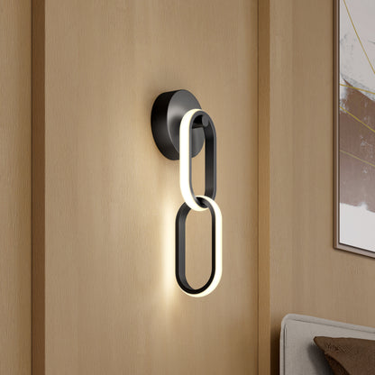 The Artika Catena modern LED wall sconce 5 CCT features two interlocking oval LED light rings mounted on a light wood panel wall, positioned next to a bed and abstract artwork.