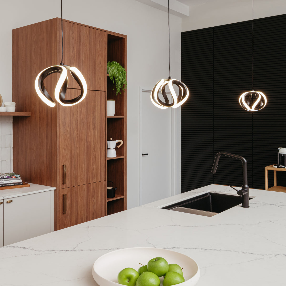 A modern kitchen with a white island, black sink, a bowl of green apples, wooden cabinets, a small potted plant, and three pendant lights with unique circular designs hanging above the island.