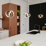 A modern kitchen with a white island, black sink, a bowl of green apples, wooden cabinets, a small potted plant, and three pendant lights with unique circular designs hanging above the island.