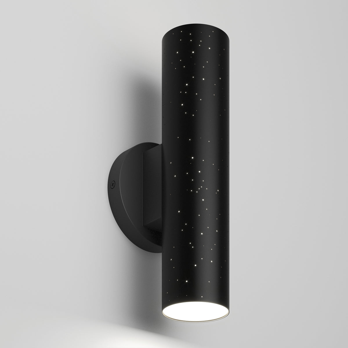 The Artika Cosmic indoor-outdoor LED wall light 3 CCT is a modern black cylindrical sconce with star-like perforations, casting light downward when mounted on a plain white wall.