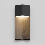 A modern wall sconce with a rectangular design, featuring a black top and bottom, wood-textured center, and a light glowing from the upper section against a light gray wall.